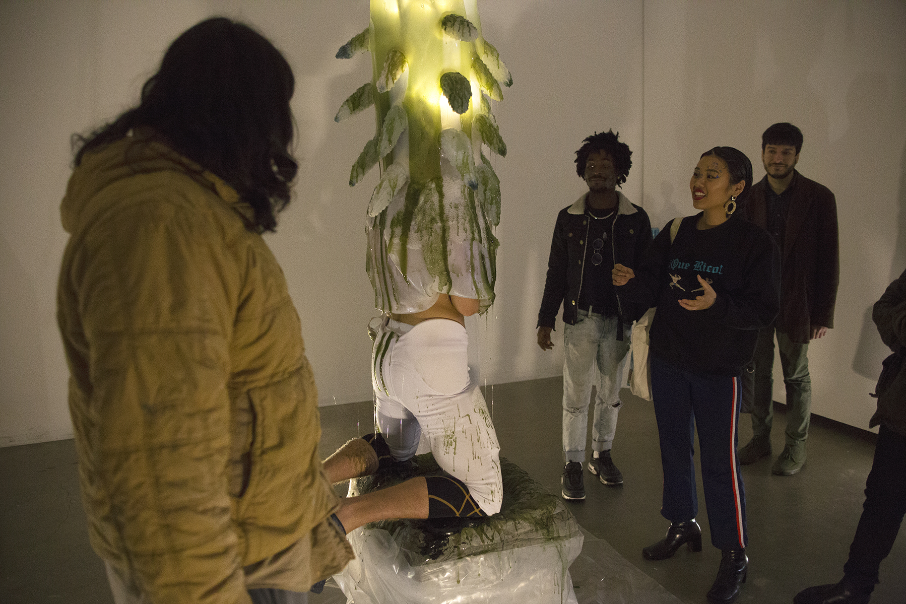 A man engulfed in a gelatenous form covered in tentacles knealing over a pool of green slime in a gallery while people watch